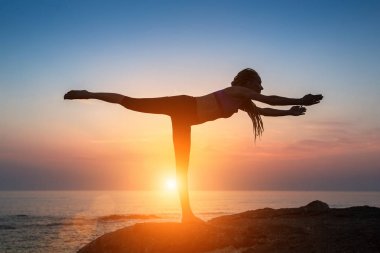 Genç kadının günbatımı sırasında deniz kıyısı jimnastik egzersizleri yapması silüeti.