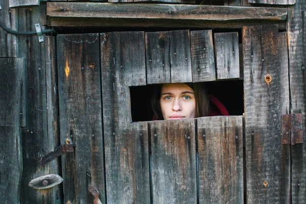Young woman looking through the crack of a locked wooden shed.