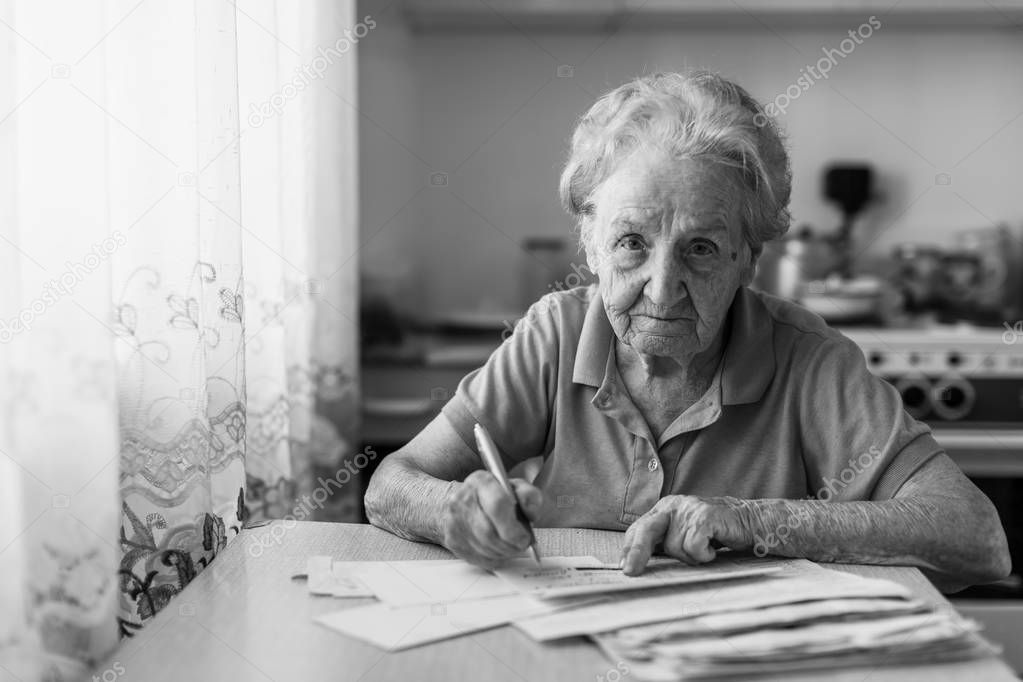 Elderly woman fills out utility bills sitting in the kitchen. Black-and-white photo.