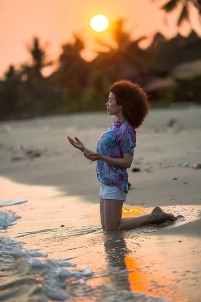 年轻的混血女子跪在热带海滩上 在一个惊人的日落 — 图库照片