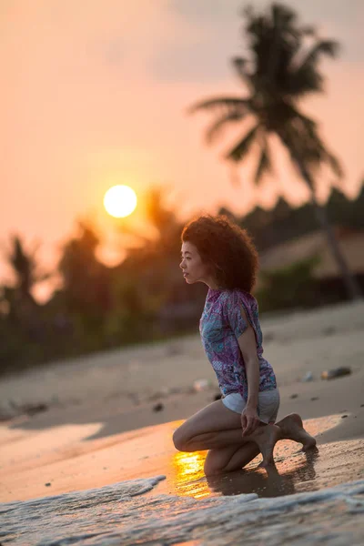 すばらしい夕日の中に熱帯のビーチの上にひざまずく若い混血女性 — ストック写真