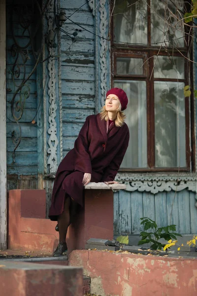 古い木造の家のポーチに座っているバーガンディコートとベレー帽の女性 — ストック写真