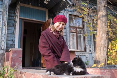 Sonbahar elbiseli bir kadın, klasik bir kır evinin verandasında kedisiyle oynuyor..