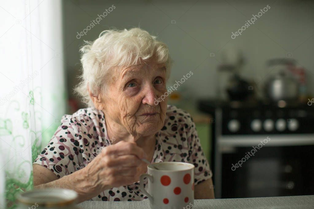 Old woman pensioner is sitting in the kitchen drinking tea. 