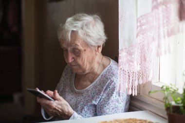 Yaşlı kadın elinde akıllı bir telefonla odada oturuyor..