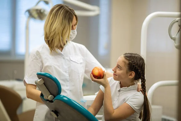 Female dentist gives apple for smiling teenage girl in dental clinic.