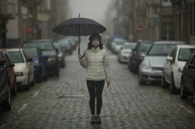 Salgın. COVID-19, Coronovirüs. Antiviral maskeli çok kültürlü bir Asyalı kadın bulutlu bir havada şemsiyelerle sokakta duruyor.. 