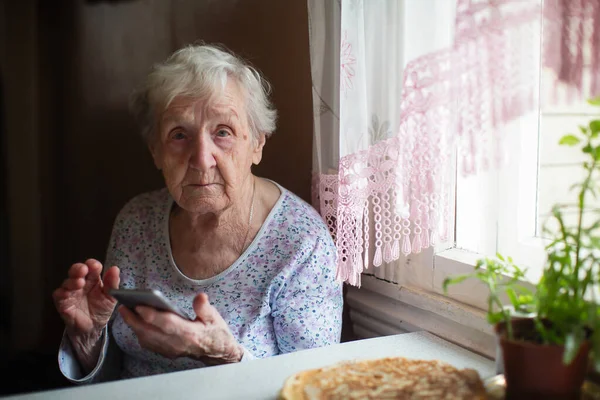 一位老年妇女养恤金领取者坐在家里 手里拿着智能手机 — 图库照片
