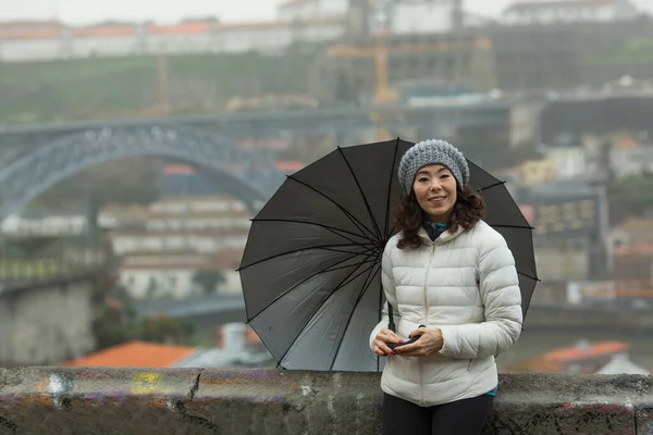 在葡萄牙波尔图的多姆路易斯一号桥的背景上拿着一把雨伞的亚洲妇女 — 图库照片