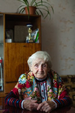 Parlak elbiseli yaşlı kadın evinde masada oturuyor..