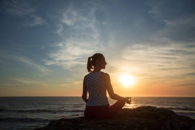 Yoga yapan kadının silueti okyanus sahilinde, gün batımında meditasyon yapıyor..  