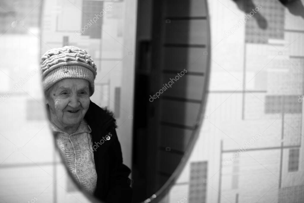 An old woman reflection in the mirror wearing winter outerwear. Black and white photo.