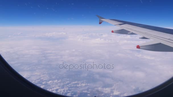 Bir uçak penceresinden görünümü: mavi gökyüzü ve bulutlar üzerinde bir uçak kanadı — Stok video