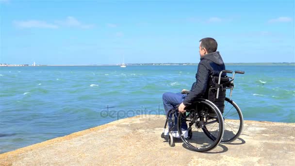 Jovem deficiente sentado em uma cadeira de rodas e olha para o mar — Vídeo de Stock