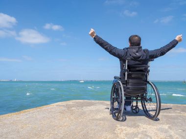 Disabled young man sitting in a wheelchair and looks at the sea  clipart
