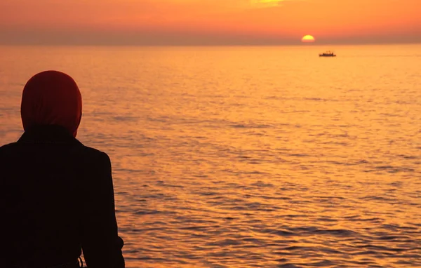 Silhouette of arab woman in traditional muslim black hijab or jilbab looking on the beautiful golden sunset in the sea and sailing ship. Back view. Peaceful serene landscape. Middle East.