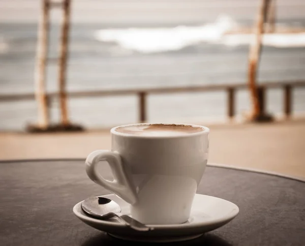 Coffee cup on round table in cafe terrace on the old pier with rusty railing and the view on the sea with waves in cold cloudy day. Loneliness idea. Aged photo.