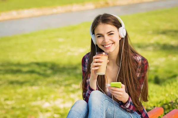 Hermosa adolescente en el parque con auriculares, teléfono inteligente y café para llevar —  Fotos de Stock