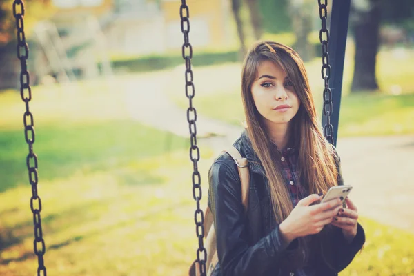 Nádherná mladá žena v parku na houpačce pomocí chytrého telefonu — Stock fotografie