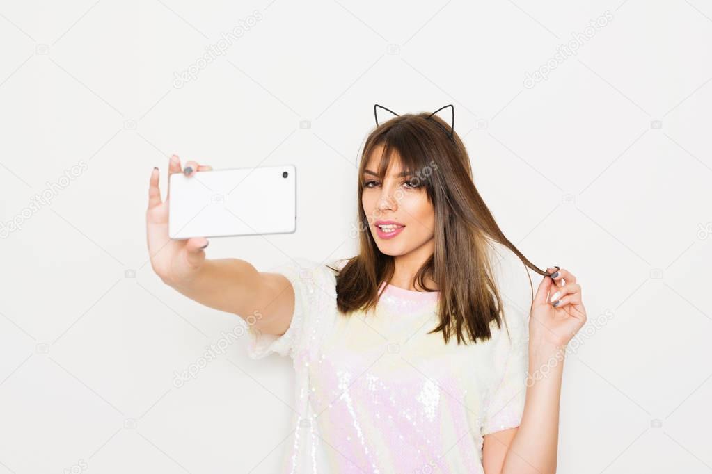 Modern young woman in sequined shirt and cat ears taking a selfie on smart phone