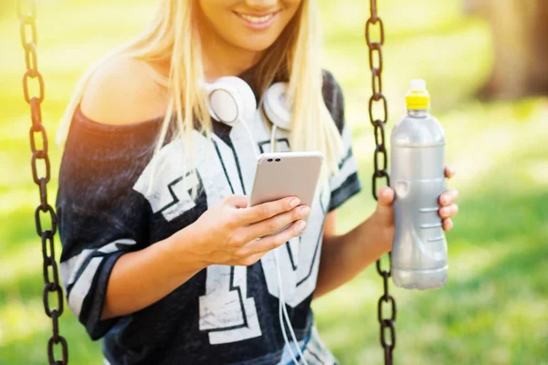 Тысячелетняя девочка-подросток в наушниках, с помощью смартфона в парке сидит на качелях, улыбается, держит бутылку воды — стоковое фото