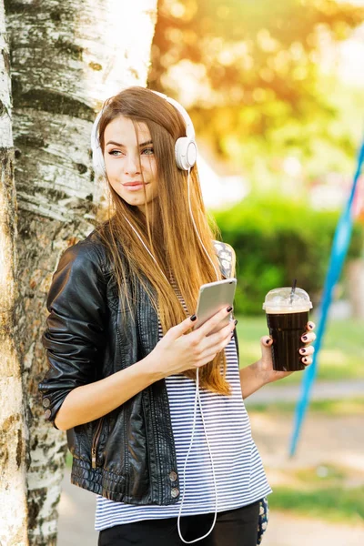 Krásné dospívající dívka s kávou, chytrý telefon a sluchátka v parku — Stock fotografie