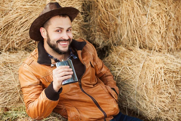 Jovem sorrindo relaxado cowboy em jaqueta de couro marrom que coloca na palha — Fotografia de Stock