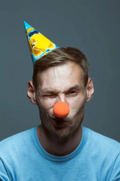 Funny young man in birthday hat with clown nose making a grimace