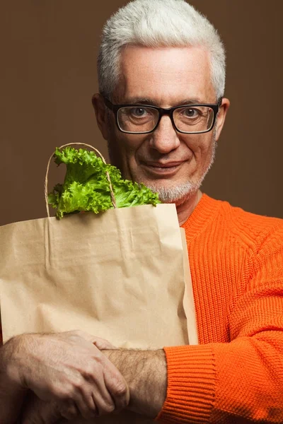 Old hipster vegan concept. Portrait of fashionable 60-year-old man in orange sweater over beige, pastel background holding paper pack with salad. Trendy haircut, glossy grey hair. Studio shot