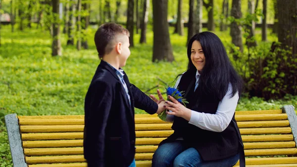 De zoon geeft zijn moeder een vers boeket van muscari blauwe bloemen op een bankje in het park. — Stockfoto