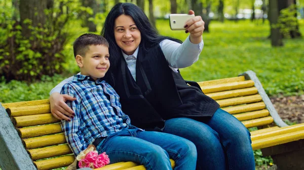 Мама и сын в парке на жёлтой скамейке фотографируются на мобильный телефон . — стоковое фото