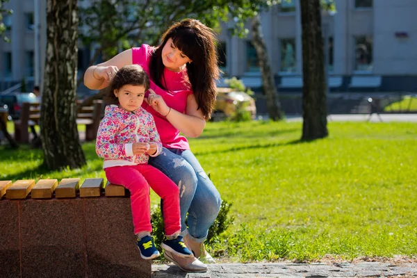 Mor gør en frisure til sin lille charmerende datter i parken på en bænk. Mors dag . - Stock-foto