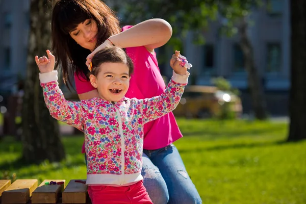 Мама робить зачіску з малою чарівною донькою в парку на лавці. День матері. — стокове фото