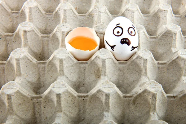 Egg with drawn cartoon faces and one broken egg in tray.