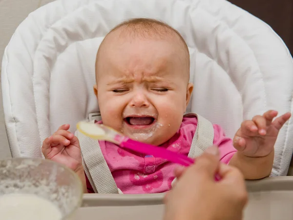Mother feeding her baby girl with a spoon. The baby is crying and does not want to eat.