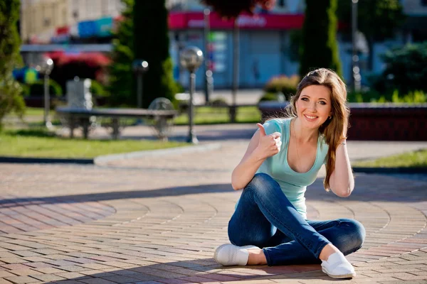 A young and pretty girl in jeans sits on a sidewalk tile in the middle of a street in the city and shows a thumbs-up.