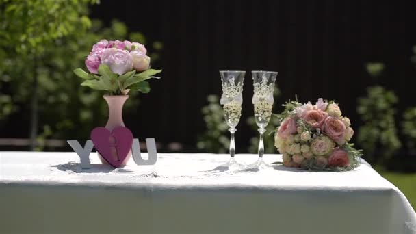 Bruidsdecor op tafel: een bruidsboeket, twee glazen champagne in de tuin. — Stockvideo