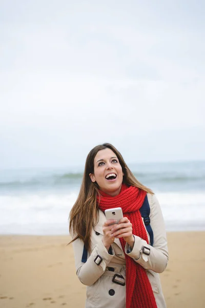 Mujer alegre mensajes de texto en el teléfono inteligente en la playa en otoño —  Fotos de Stock