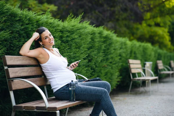 Donna incinta seduta in una panchina del parco ad ascoltare musica — Foto Stock