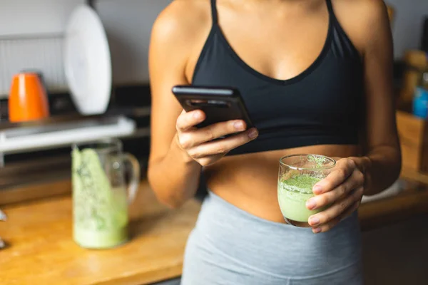 健康饮食的概念 一个身材苗条的女人拿着一个绿色的排毒软糖 在厨房的智能手机上使用营养应用程序 合适的早餐 — 图库照片