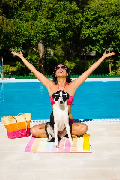 水泳プールで夏に一緒に幸せな女性とかわいい犬の日光浴 美しい女の子と彼女のペット身に着けているサングラスと楽しい休日にプールサイド — ストック写真