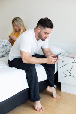 Social networks addiction and relationship problems concept. Man and woman in bed ignoring to each other for checking their messages on their phones. clipart