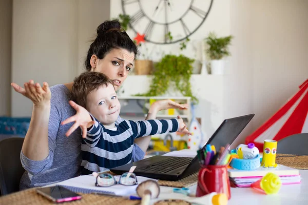 Busy mother working from home and taking care of her child. Stressed woman telecommuting on laptop computer with her son.