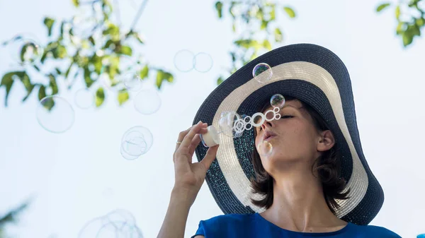 Woman wearing blue dress with hat blowing bubbles — Stock Photo, Image