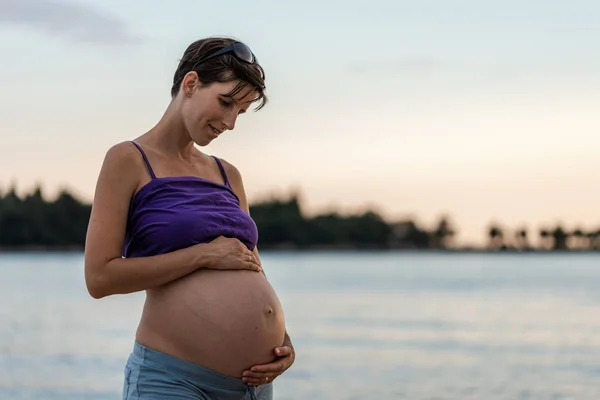 Mladá těhotná žena stojící za soumraku u moře — Stock fotografie