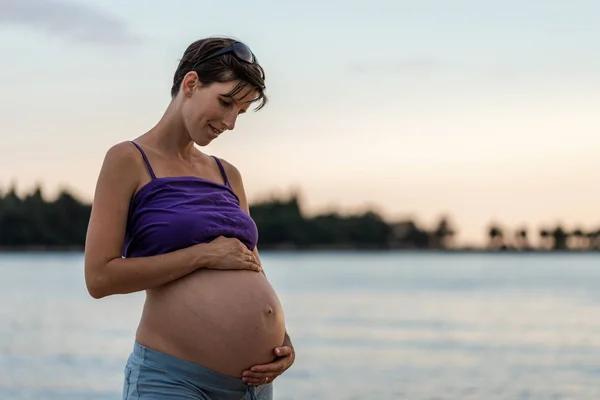 Ung gravid kvinna som står vid havet i skymningen Stockbild