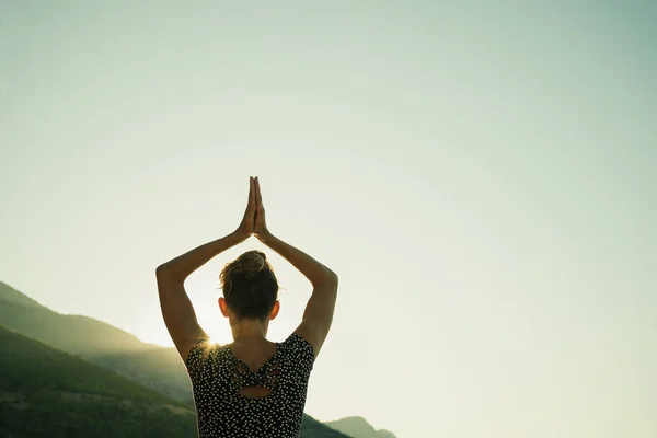 View from behind of a young woman making an assana pose with her arms joined above her head as she does her morning yoga workout at sunrise.