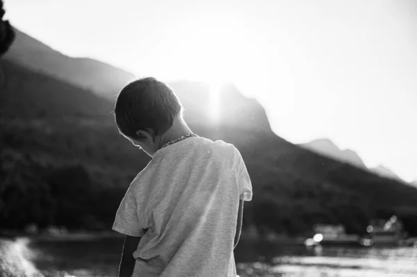 从后面看 一个小男孩站在海边 太阳从山后升起 单色图像 — 图库照片
