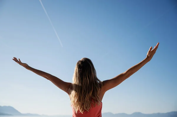 View from behind of a young woman with long hair standing  outside under clear summer sky with her arms raised and spread.