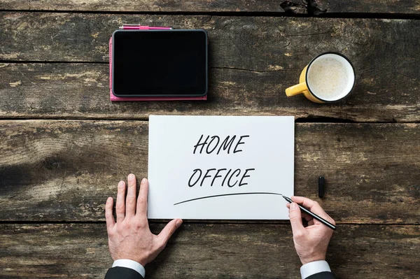 Top view of businessman writing a Home office sign on white paper with digital tablet and cup of coffee placed next to it.